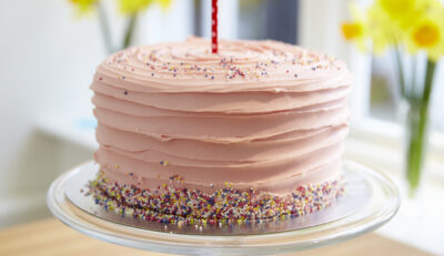 pink cake with one candle on it in front of daffodils