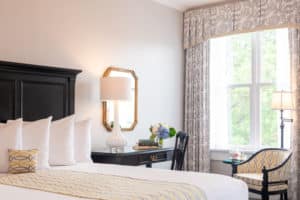 King bed with cherry headboard, cherry desk with lamp beside bed by window with gray drapes and cushioned chair