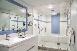 Glass enclosed tiled shower / tub combination, white vanity with double sinks, blue framed mirror above.