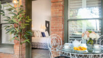 View through door into bedroom from terrace of red brick building, round glass table set for breakfast, potted plant by door