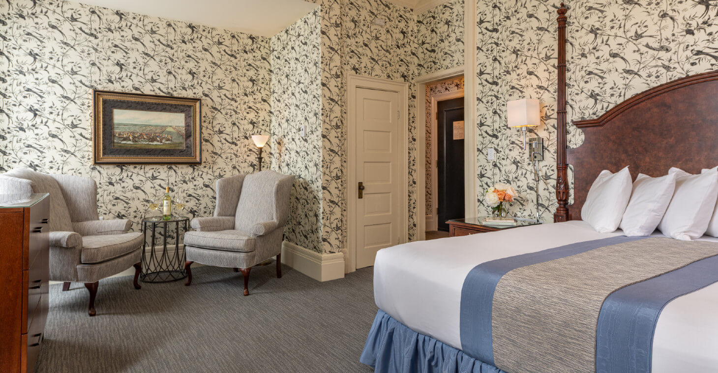 Wallpaper with birds in King bedroom with two-poster bed, grey queene anne chairs in nook with lamp, stamp and art hanging on wall.