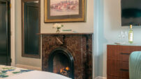 Close-up view of fireplace with frame art hanging over mantel, full-length mirror on wall, blue chair by dresser under tv on wall