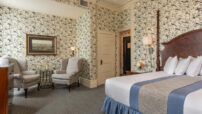 Wallpaper with birds in King bedroom with two-poster bed, grey queene anne chairs in nook with lamp, stamp and art hanging on wall.