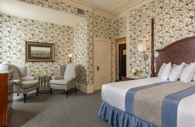 Wallpaper with birds in King bedroom with two-poster bed, grey queene anne chairs in nook with lamp, stamp and art hanging on wall.