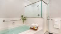 Two-person whirlpool tub with grab bar, tray with roll towels on edge, tiled glass enclosed shower