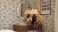 Close-up view of desk in corner with lamp and flowers in King bedroom with birds on wallpaper under oval mirror