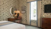 Wallpaper with birds in King bedroom with desk in corner under oval mirror, dresser under flat screen tv by window