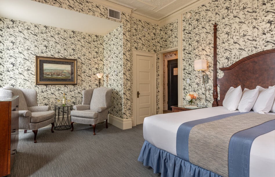 Wallpaper with birds in King bedroom with two-poster bed, grey queene anne chairs in nook with lamp, stamp and art hanging on wall.