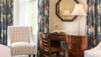 Oak desk with opened book and wine glass in between windows with floral drapes and checkered chairs