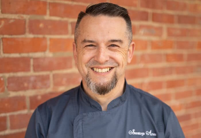 Headshot of smiling chef Tim by red brick building wearing grey Saratoga Arms shirt