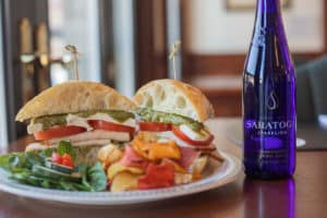 White plate with sandwich with toothpicks, salad and pita chips, blue bottle with Saratoga sparking water