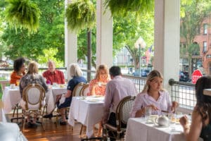 Long covered porch by street with linen covered tables and happy guests visiting at breakfast