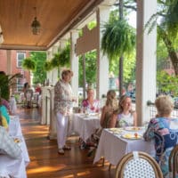 Long covered porch by street set for breakfast with hostess mingling with guests