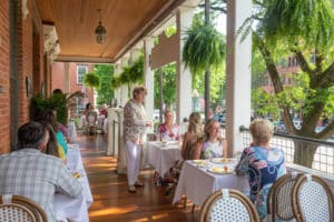 Long covered porch by street set for breakfast with hostess mingling with guests