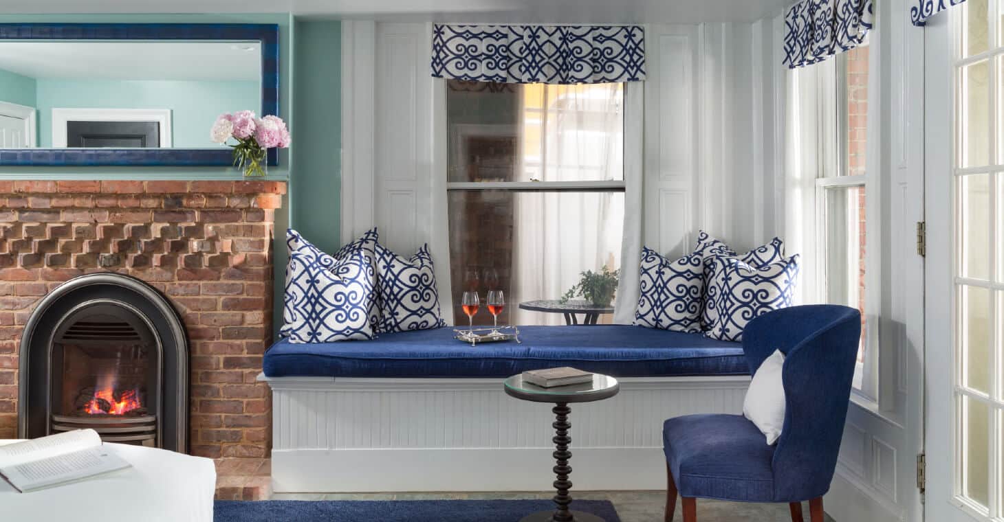Brick fireplace with blue framed mirror hanging above, blue cushioned bench by window with 2 red wine glasses