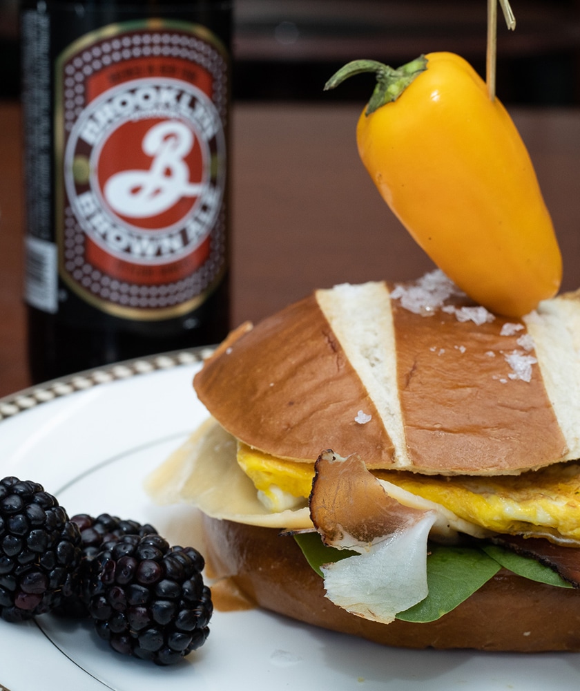 A bottle of Brooklyn brown ale beer next to a large sandwich with egg, cheese, and bacon on a bun. There is a yellow pepper skewer through the top of the sandwich and some blackberries scattered around the plate. 