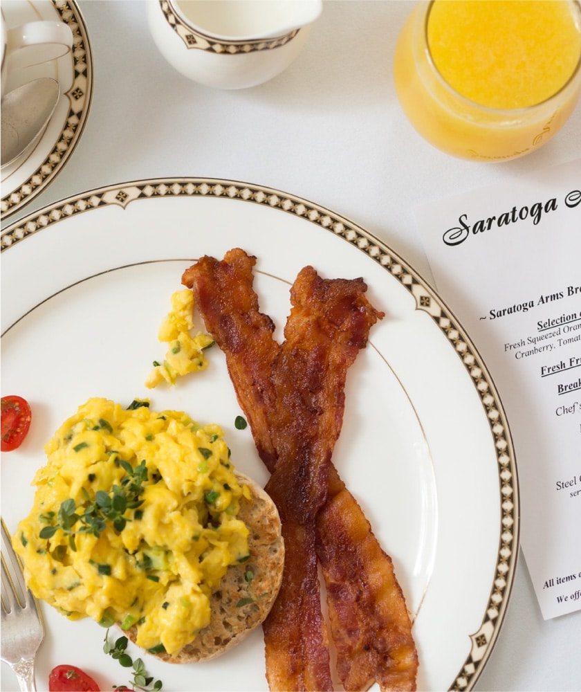 A top-down shot of a large plate of bacon and eggs sprinkled with seasoning and a few cherry tomatoes. There is a cup of orange juice and a menu to the right of the plate.