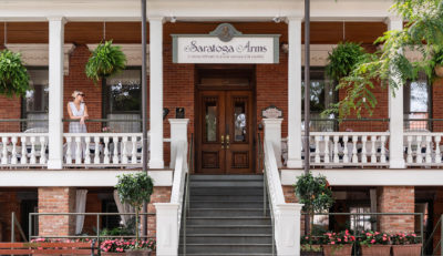 Front entrance to the Saratoga Arms Hotel with a large staircase leading to a brick building decorated with hanging plants. A woman is standing to the left of the entrance looking into the distance.