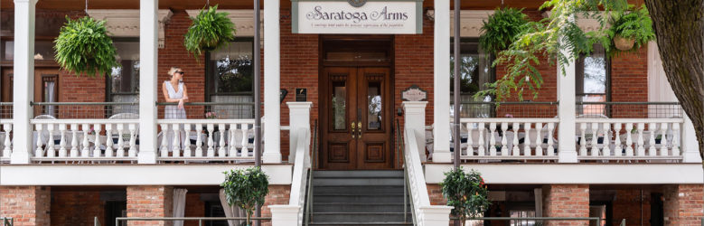 Front entrance to the Saratoga Arms Hotel with a large staircase leading to a brick building decorated with hanging plants. A woman is standing to the left of the entrance looking into the distance.
