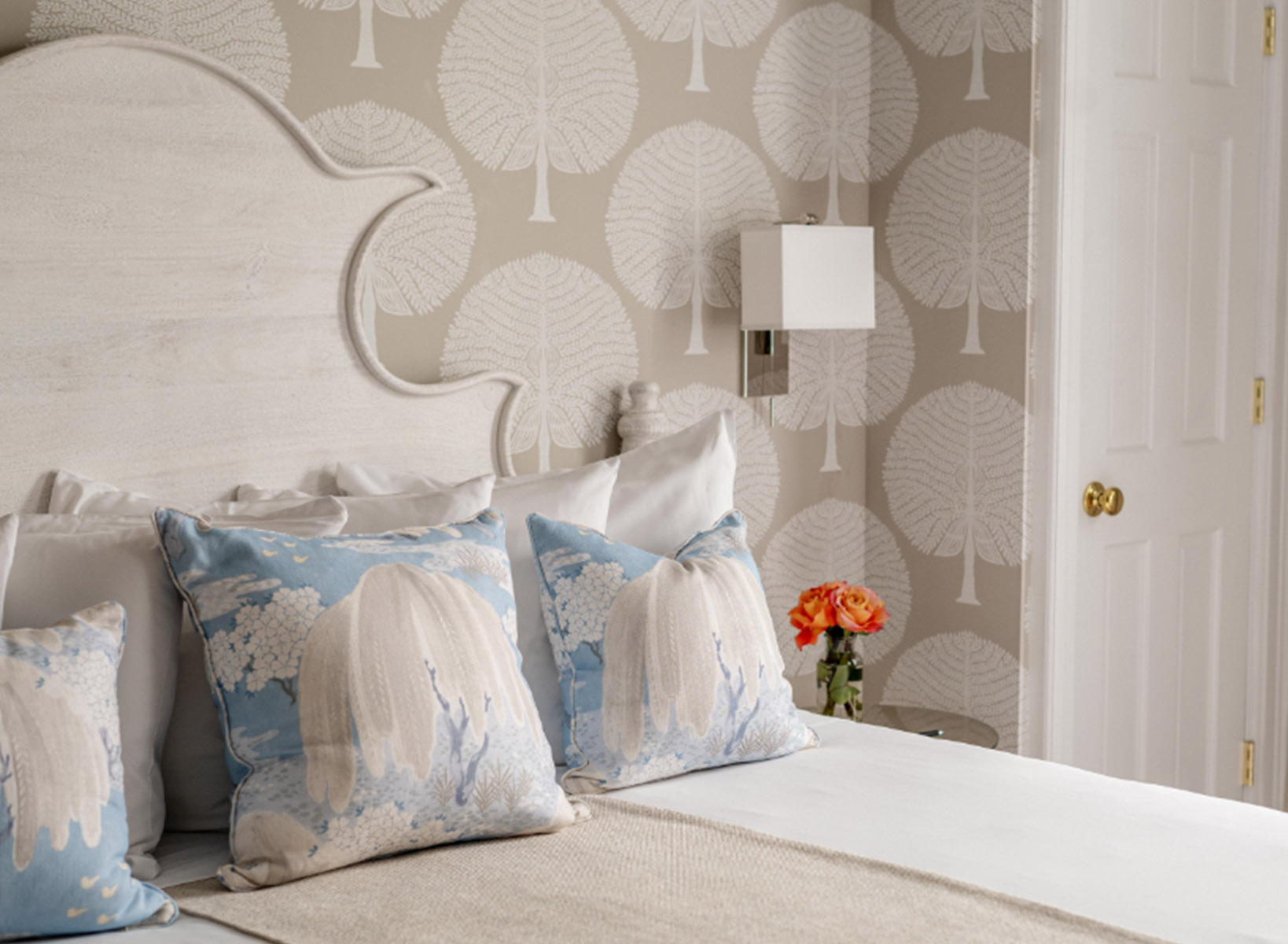 View of a bed's headboard with many white fluffy pillows and 3 decorative blue pillows on top of them. There is a vase with orange roses next to the bed on the side-table. 