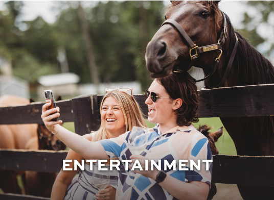 2 women take a selfie in front of a large brown horse at the racetrack.