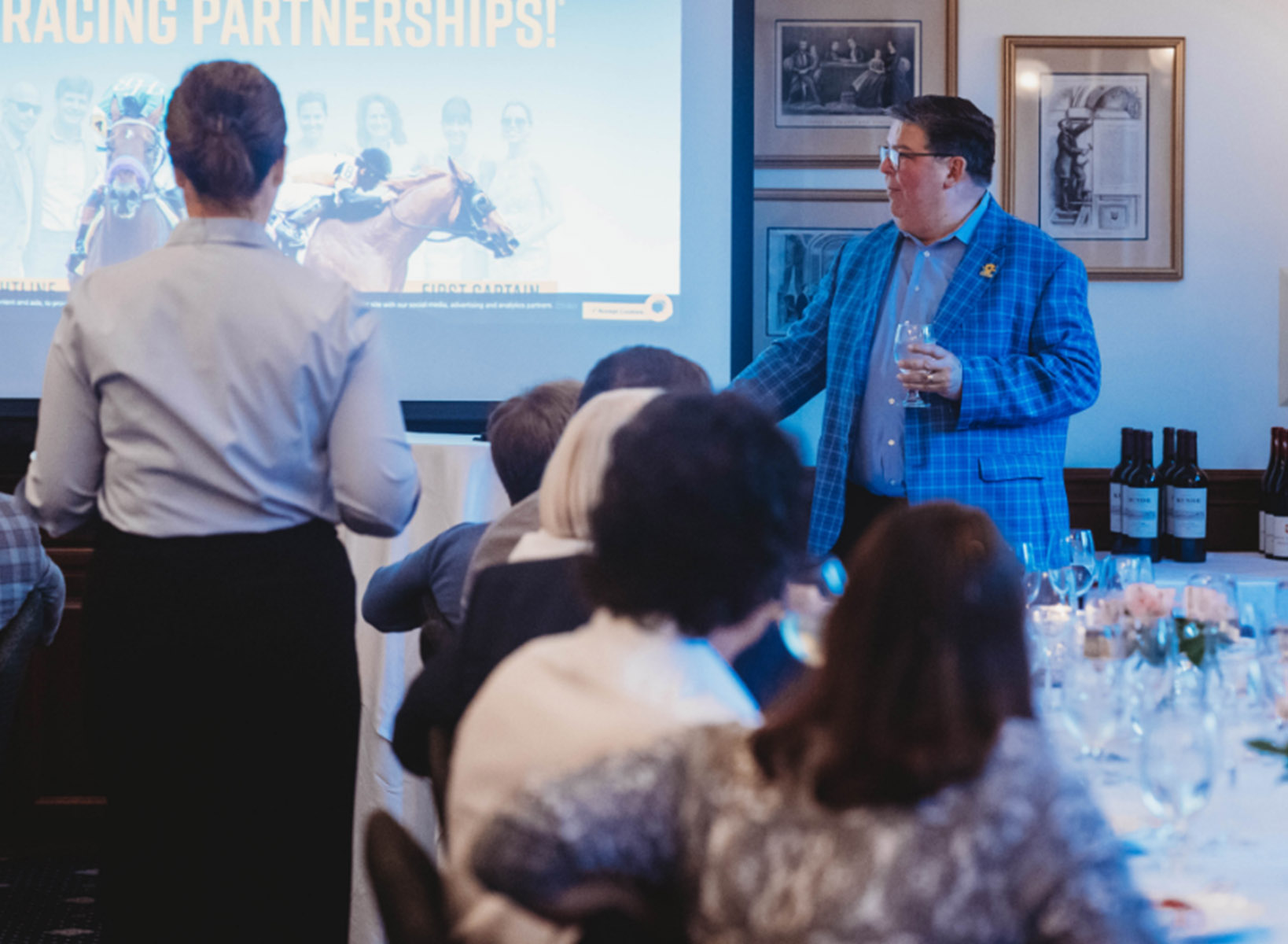 A group of business employees sitting at a long table drinking wine while looking at a projector displaying racing horses and the text Racing Partnerships on the display. A man is standing next to the projector wearing a blue suit with a wine glass in his hand.