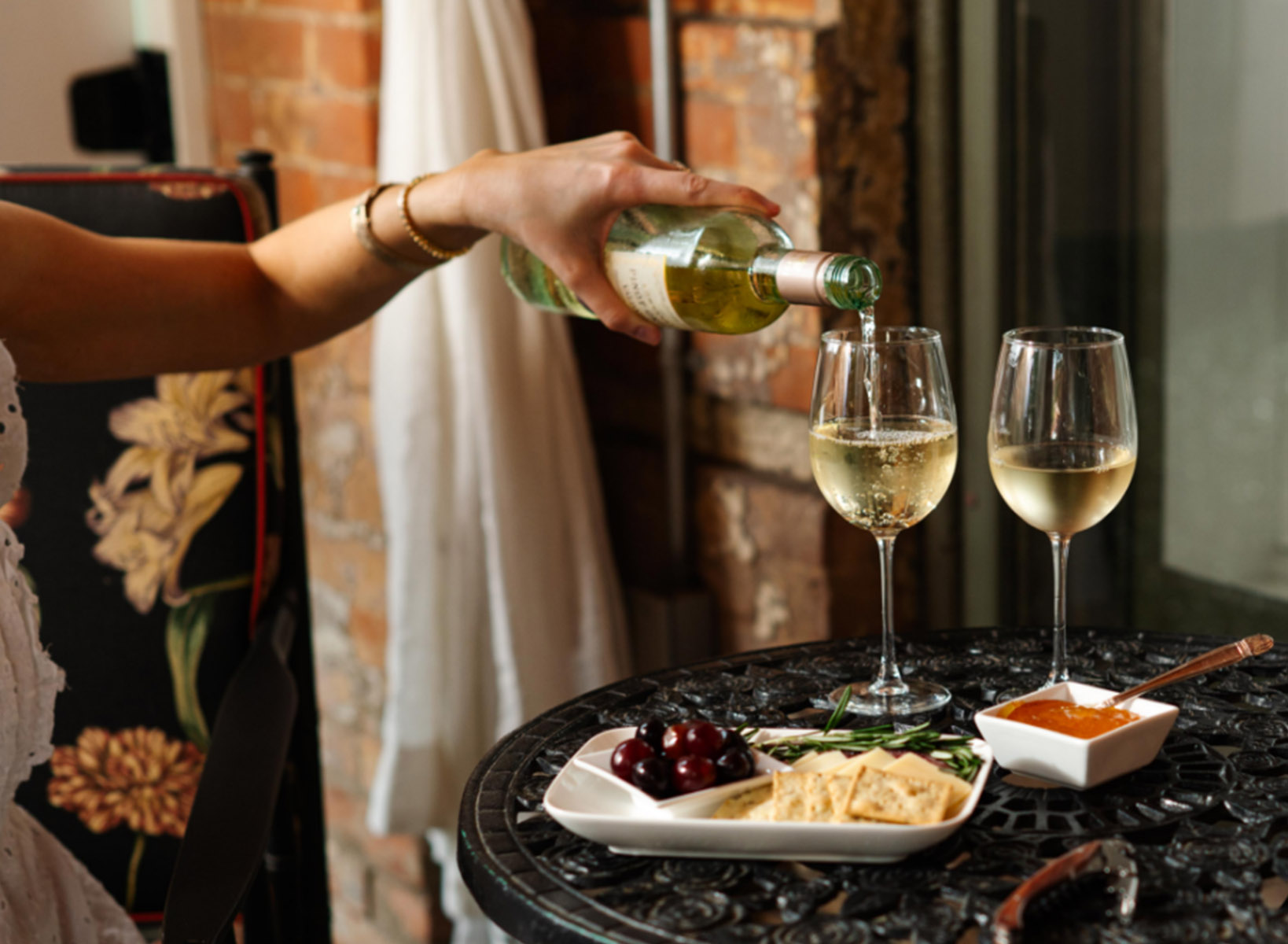 A hand pouring a glass of white wine into 2 wine glasses against a brick background. On the table with the wine glasses is a spread of crackers, cherries, and jam.