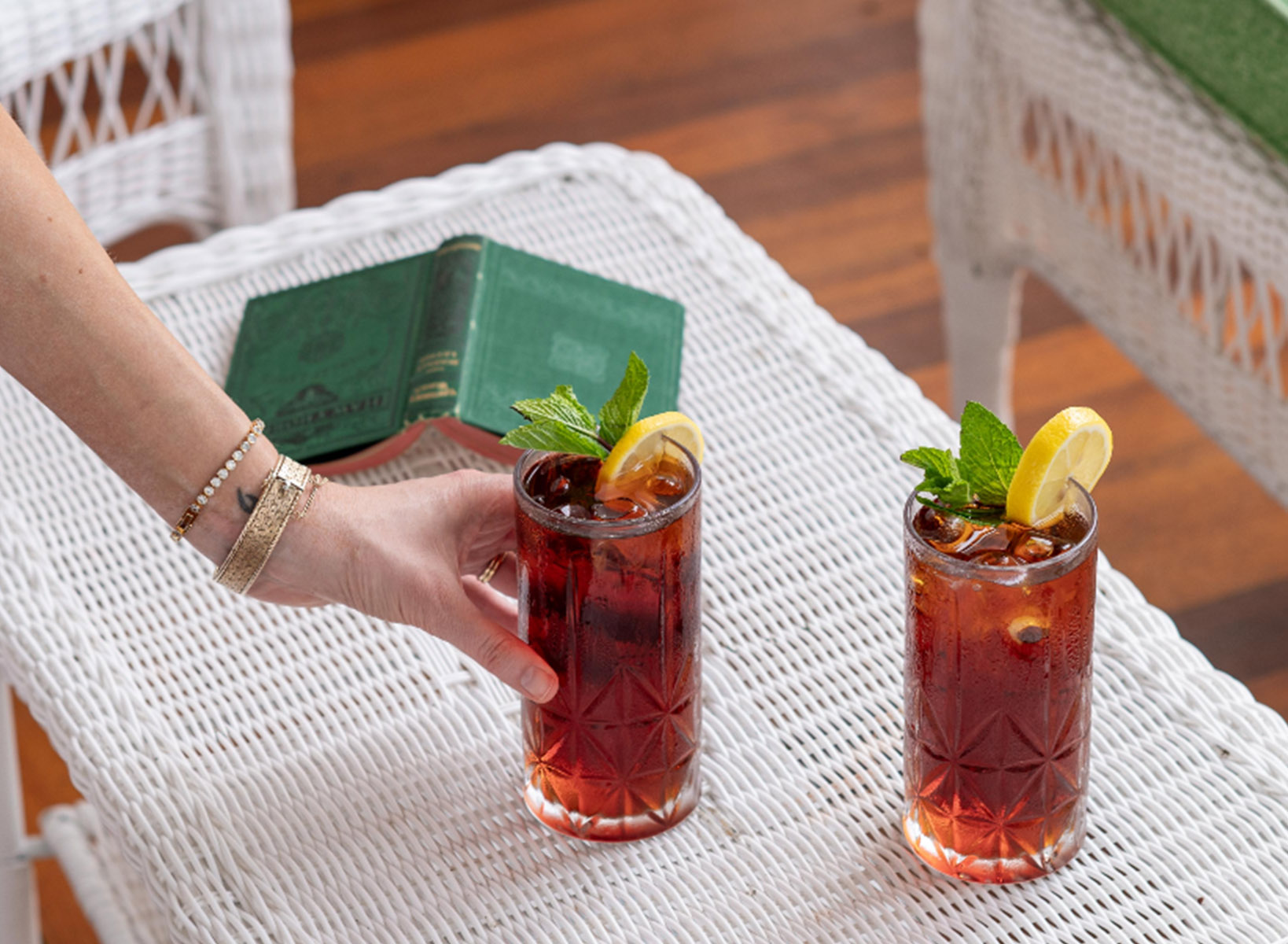 A feminine hand with bracelets grabbing a tall glass of tea with a lemon and mint on the rim of the glass. There is a green book opened in the background. 