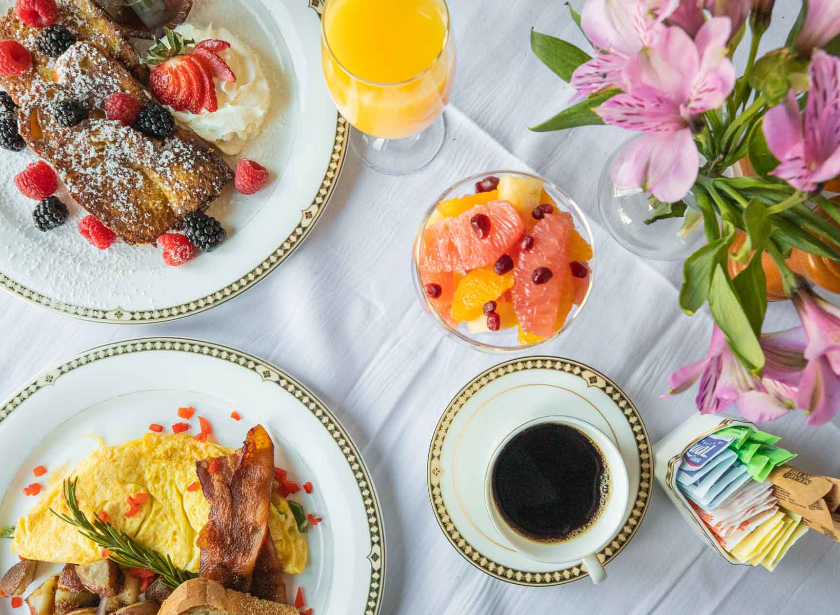 Overhead shot of a full breakfast with coffee, fresh fruit bowl, a plate of eggs and bacon, and a plate of french toast adorned with raspberries and blackberries. There is a vase of fresh pink flowers in water for decoration and a small dish of sugar for the coffee.