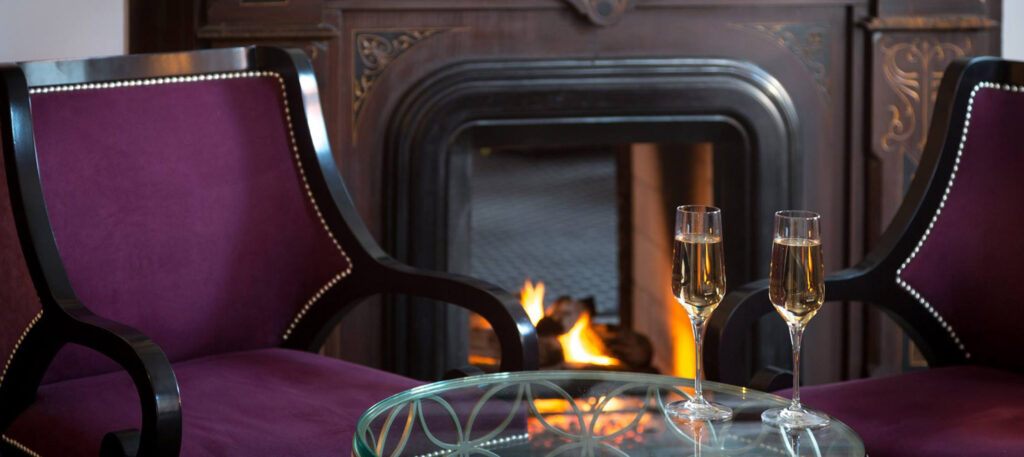 2 Cushioned velvet seats with wooden accents sit in front of a large ornate fireplace. Between the seats is a glass table with 2 long glasses of champaign.